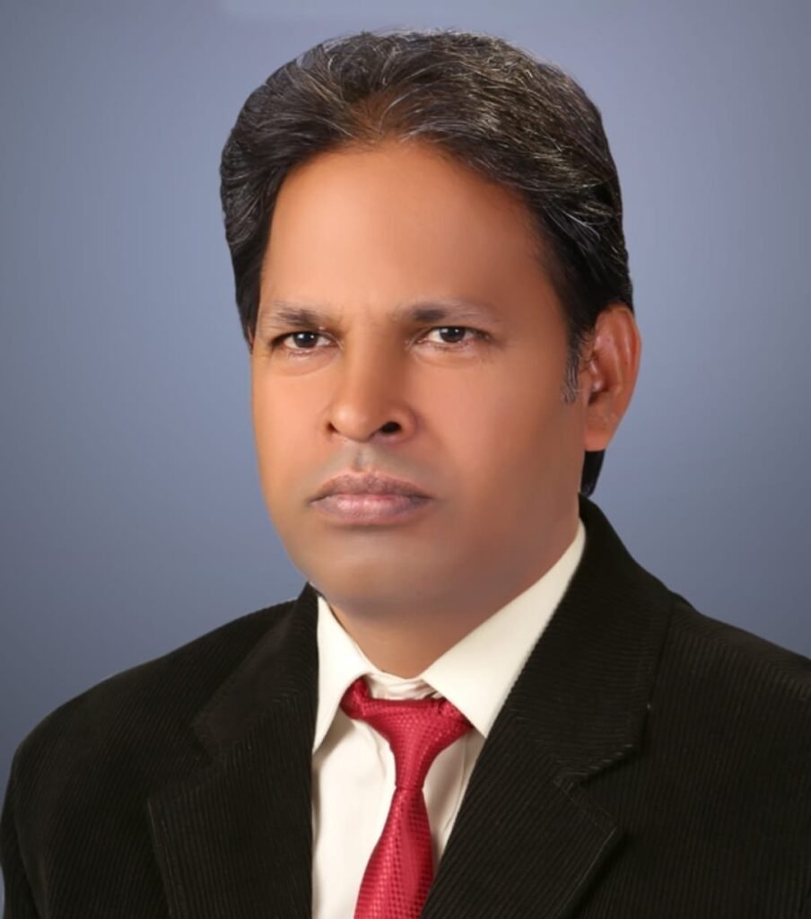 🗒️ Image Alt TextA formal studio portrait of Ashok Kumar Yadav Sir, Chemistry HOD with over 25 years of experience. He is a middle-aged man with short, styled dark hair with subtle grey highlights, looking directly forward with a serious expression. He wears a professional black textured suit blazer over a crisp white collared dress shirt, paired with a solid red tie. The background is a clean, neutral gradient of light blue and grey tones.